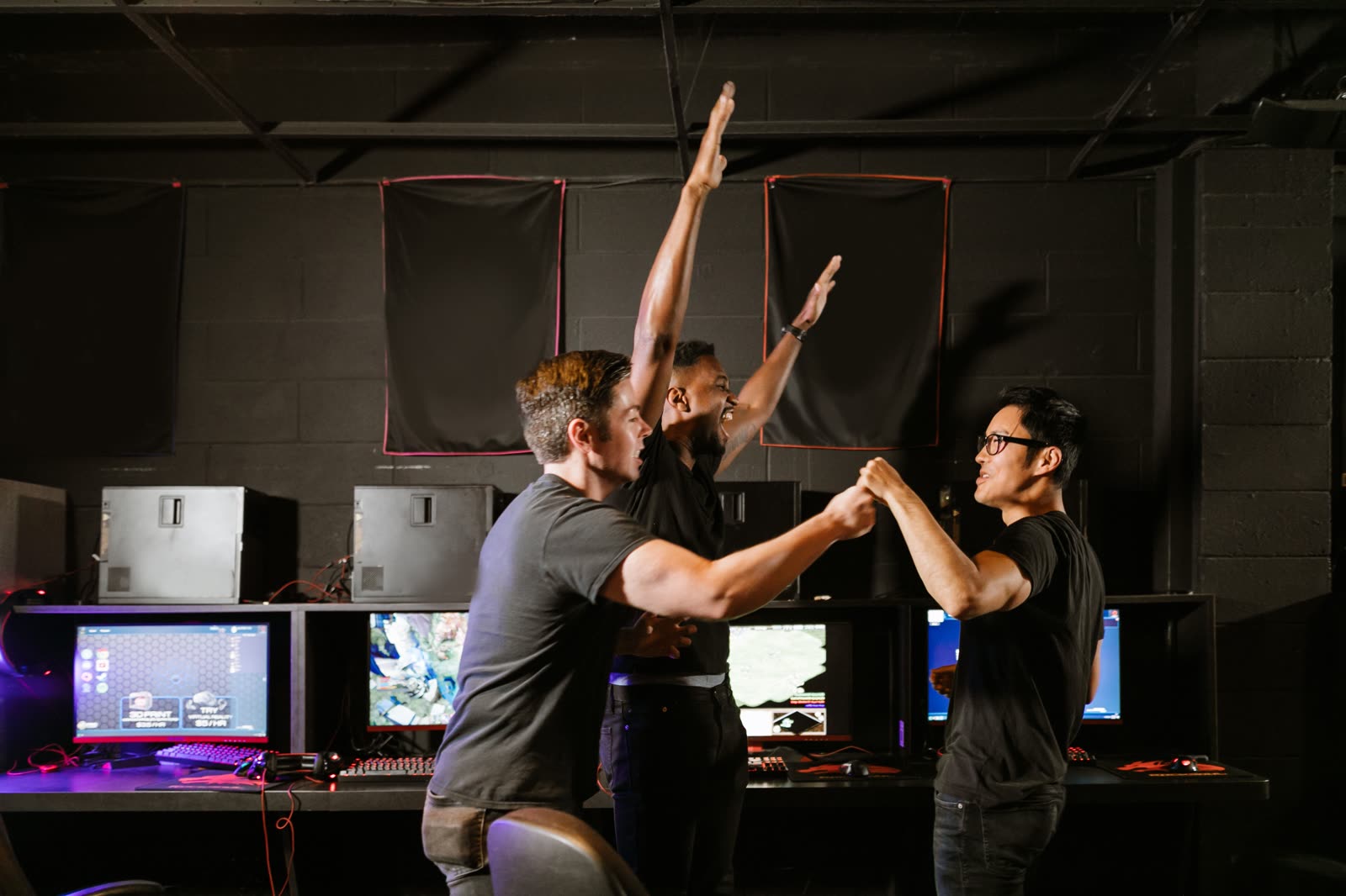 A wide dark esports room with a long lit desk of monitors as three players celebrate with raised hands